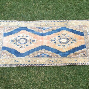 May include: A vintage rug with a blue, pink, and beige geometric pattern. The rug has a fringed edge and is laid out on a green grassy surface.