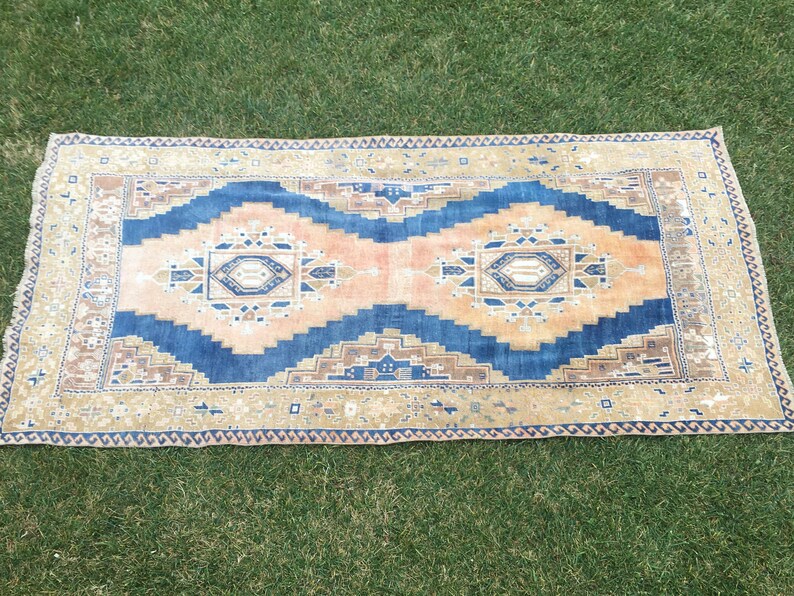 May include: A vintage rug with a blue, pink, and beige geometric pattern. The rug has a fringed edge and is laid out on a green grassy surface.