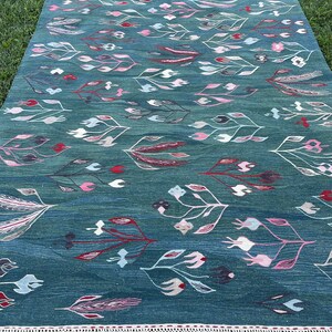 May include: A teal blue rug with a geometric pattern of stylized flowers and leaves in shades of pink, red, and white. The rug has a white fringe border.