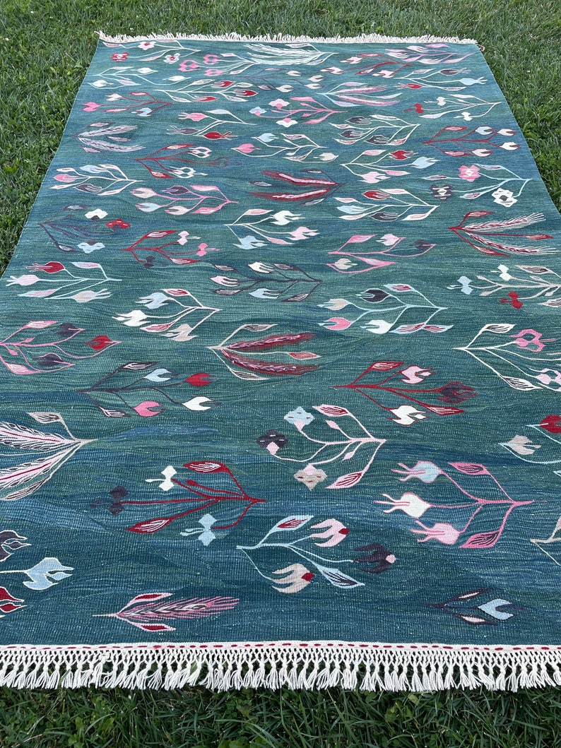May include: A teal blue rug with a geometric pattern of stylized flowers and leaves in shades of pink, red, and white. The rug has a white fringe border.