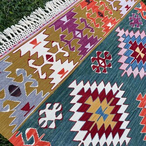 May include: A colourful rug with a geometric pattern in shades of red, orange, yellow, blue, green, purple, and white. The rug has a fringed border.