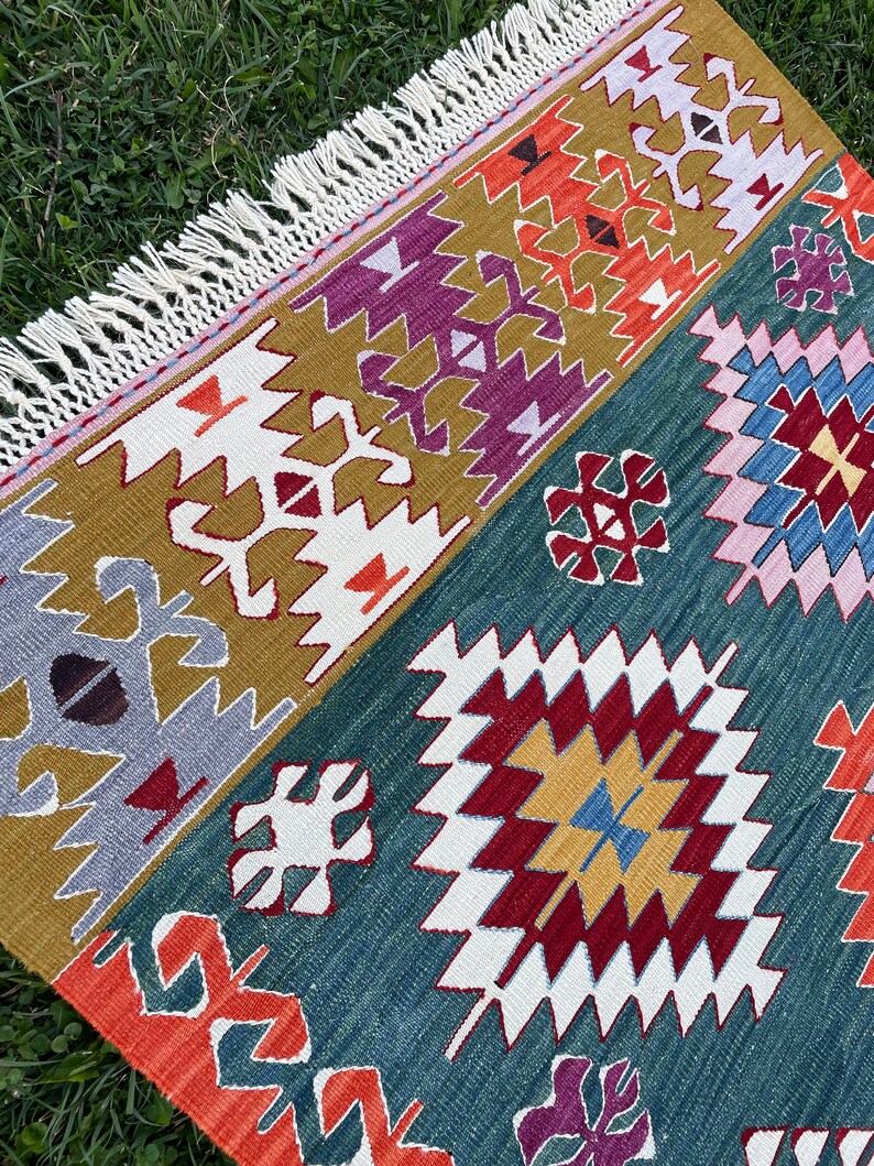 May include: A colourful rug with a geometric pattern in shades of red, orange, yellow, blue, green, purple, and white. The rug has a fringed border.