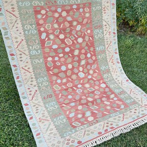 K&ouml;nnte beinhalten: Ein rot, gr&uuml;n und beige gemusterter Teppich mit Fransenbord&uuml;re. Der Teppich hat ein geometrisches Muster mit einem zentralen Medaillon.