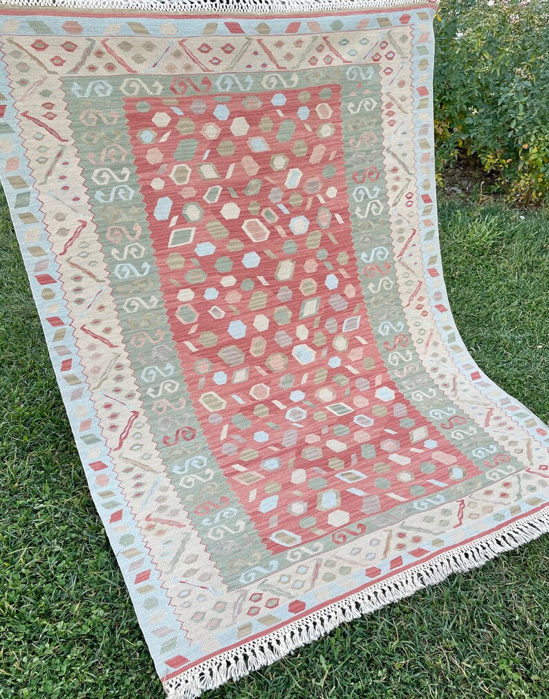 K&ouml;nnte beinhalten: Ein rot, gr&uuml;n und beige gemusterter Teppich mit Fransenbord&uuml;re. Der Teppich hat ein geometrisches Muster mit einem zentralen Medaillon.