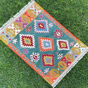 May include: A colourful rug with a geometric pattern in shades of red, orange, yellow, blue, green, and white. The rug has a fringed border.