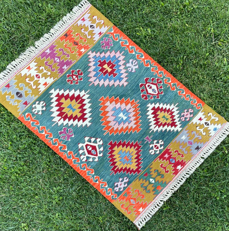 May include: A colourful rug with a geometric pattern in shades of red, orange, yellow, blue, green, and white. The rug has a fringed border.