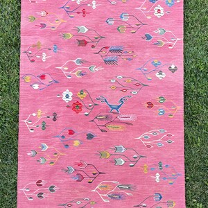 May include: A pink rug with a white fringe border features a repeating pattern of stylized floral designs in various colors, including red, blue, yellow, and green. A stylized animal figure is woven into the center of the rug.