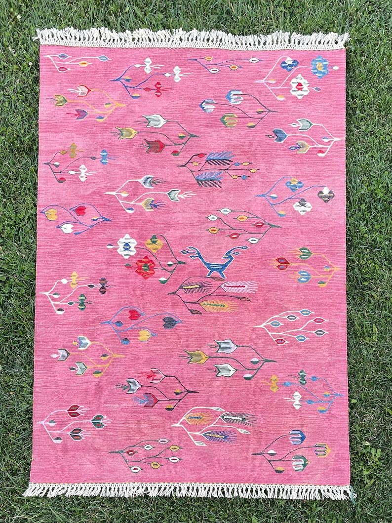 May include: A pink rug with a white fringe border features a repeating pattern of stylized floral designs in various colors, including red, blue, yellow, and green. A stylized animal figure is woven into the center of the rug.