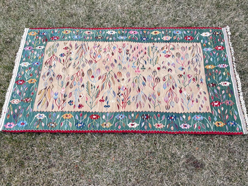 May include: A rectangular rug with a cream background and a green border. The rug features a floral pattern with pink, red, and blue accents.