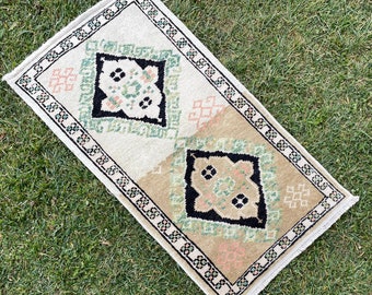 May include: A small, rectangular rug with a white background and a geometric pattern in shades of green, black, and pink. The rug has a fringed edge.