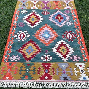 May include: A colourful, geometric rug with a green background and intricate patterns in shades of red, orange, yellow, blue, and white. The rug has a fringe border.
