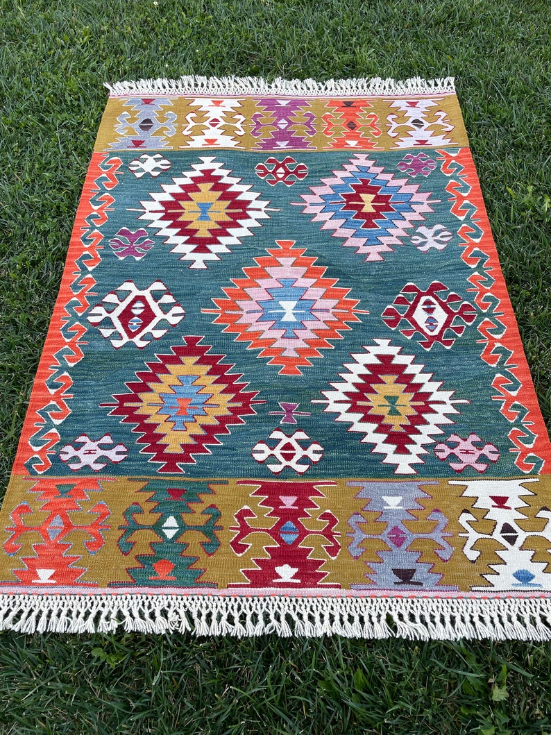 May include: A colourful, geometric rug with a green background and intricate patterns in shades of red, orange, yellow, blue, and white. The rug has a fringe border.