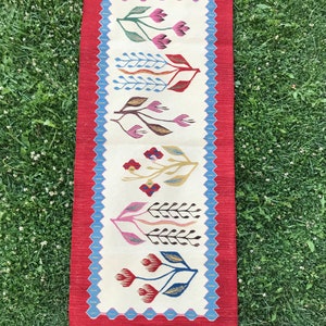 May include: A long, narrow rug with a white background and a red border. The rug features a repeating pattern of stylized flowers in shades of pink, red, blue, green, and brown.