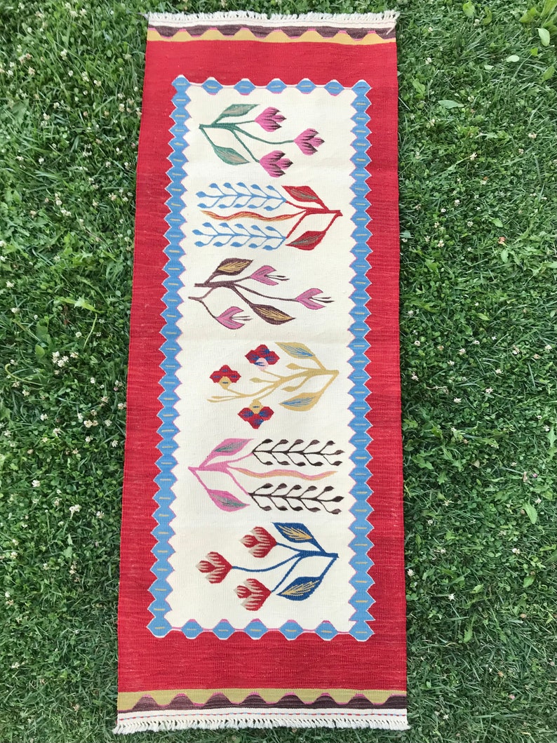 May include: A long, narrow rug with a white background and a red border. The rug features a repeating pattern of stylized flowers in shades of pink, red, blue, green, and brown.