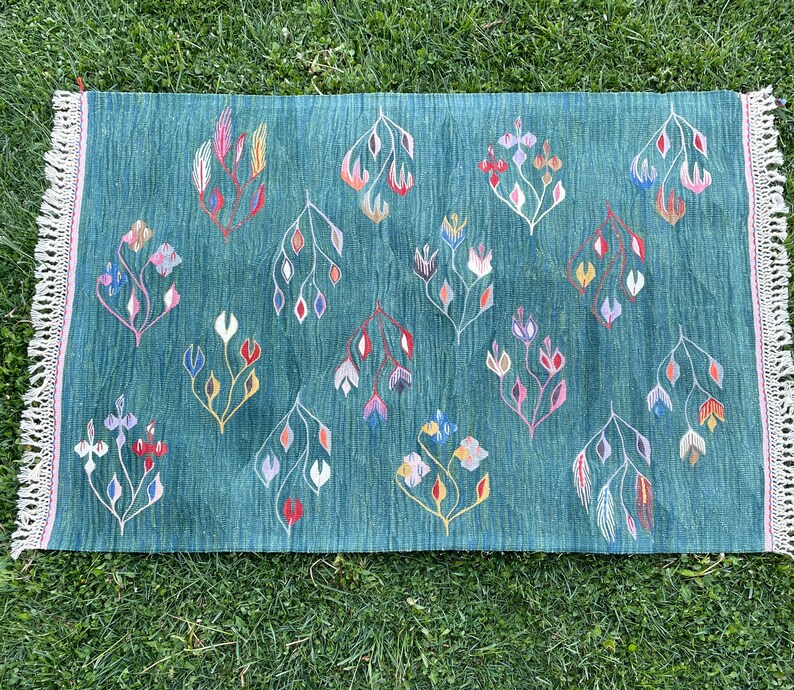May include: A teal blue rug with a fringed border features a repeating pattern of stylized floral motifs in shades of pink, red, white, and green.