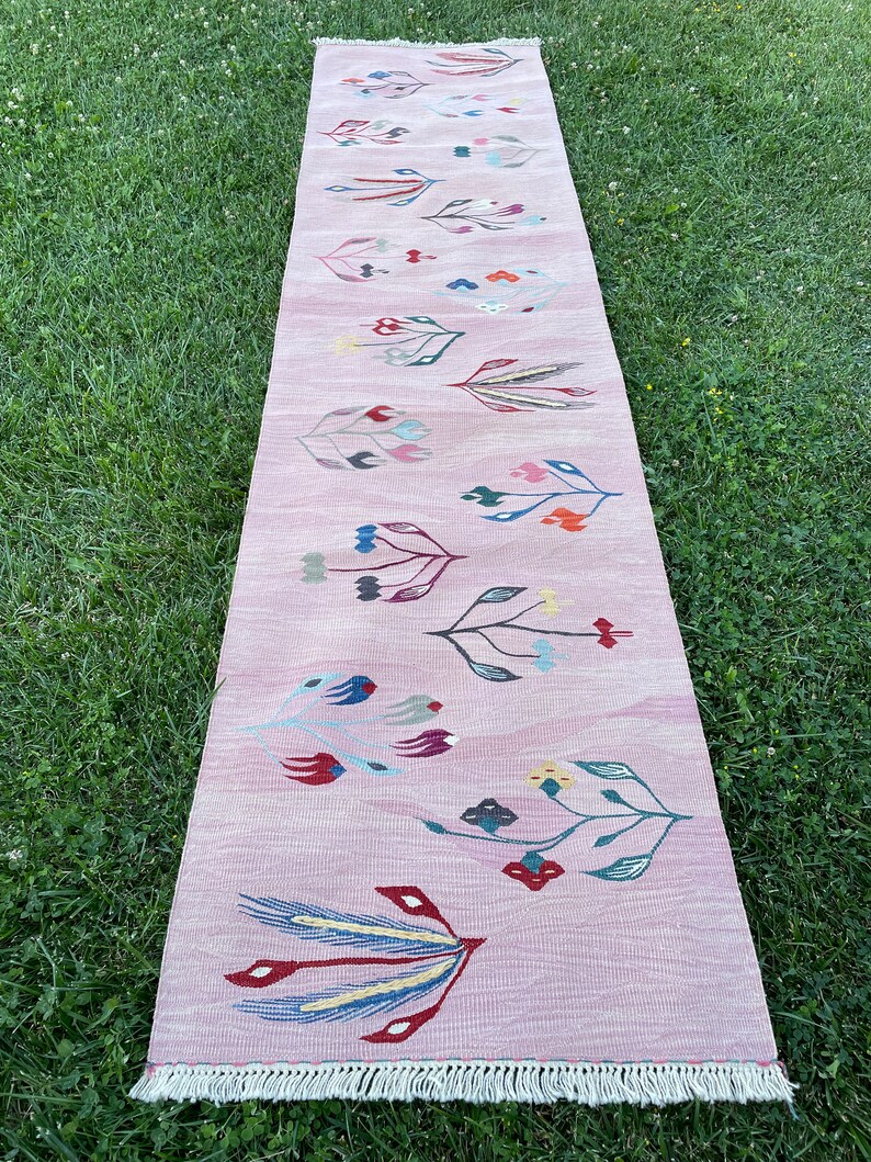 May include: A long pink rug with a floral pattern in shades of red, blue, green, and yellow. The rug has a white fringe on the edges.