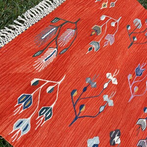 Pu&ograve; includere: Un tappeto rosso con un bordo a frangia presenta un motivo ripetitivo di disegni floreali stilizzati in tonalit&agrave; di blu, verde, bianco e giallo.