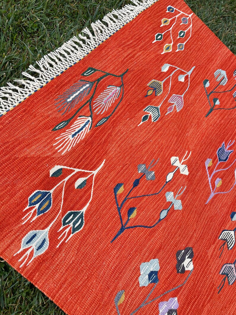 Pu&ograve; includere: Un tappeto rosso con un bordo a frangia presenta un motivo ripetitivo di disegni floreali stilizzati in tonalit&agrave; di blu, verde, bianco e giallo.