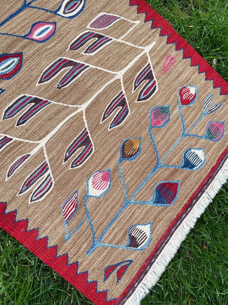 May include: A brown rug with a white, blue, and red floral pattern. The rug has a red border with a white fringe.