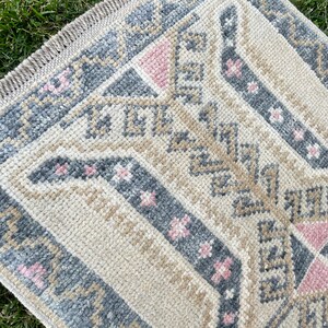 Puede incluir: Una alfombra beige con un patr&oacute;n geom&eacute;trico en tonos grises, rosa y marr&oacute;n. La alfombra tiene un borde de flecos.