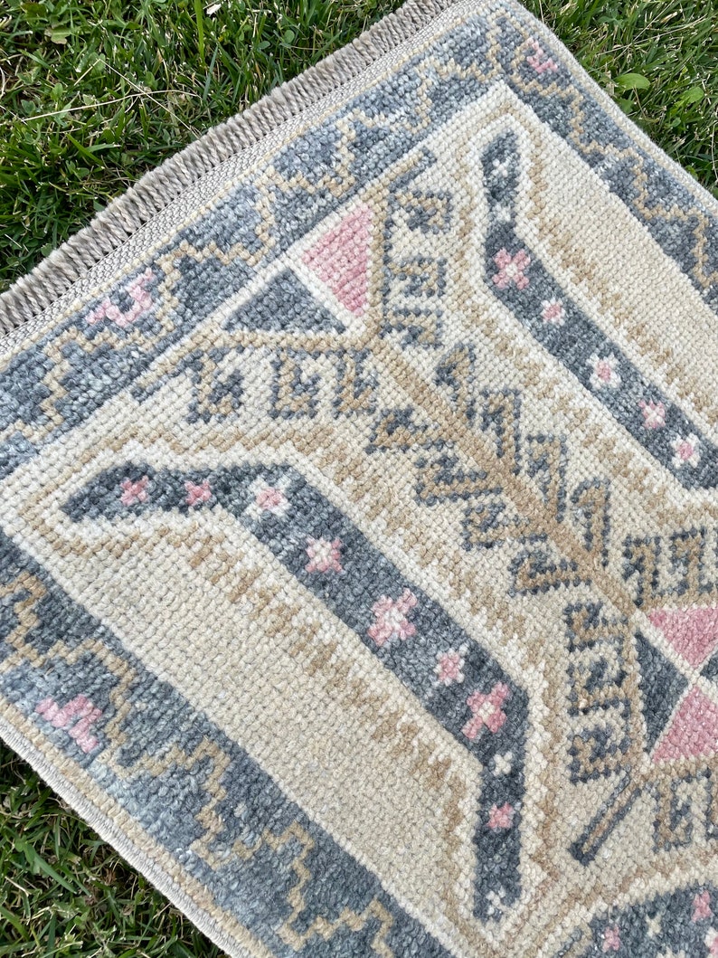Puede incluir: Una alfombra beige con un patr&oacute;n geom&eacute;trico en tonos grises, rosa y marr&oacute;n. La alfombra tiene un borde de flecos.