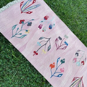May include: A pink rug with a floral design. The rug has a fringe border and features colorful flowers and leaves.