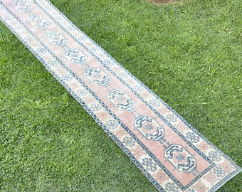 May include: A long, narrow rug with a pink background and a geometric pattern in shades of blue and white. The rug is laid out on a green grassy surface.