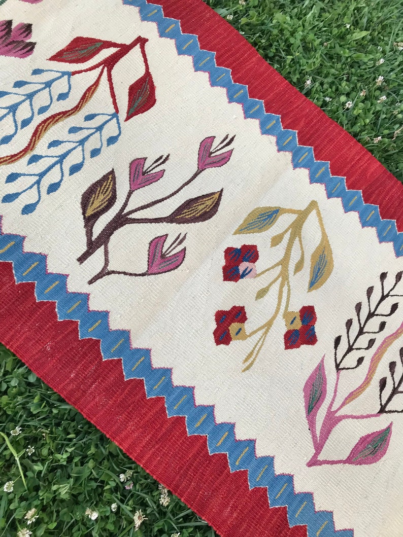 May include: A white rug with a red border and a blue and yellow geometric pattern. The rug features a floral design with pink, red, yellow, and brown flowers.