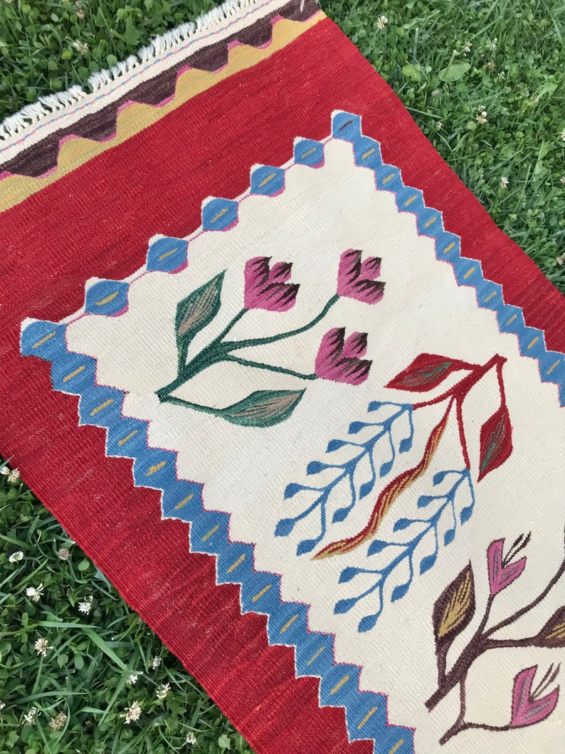 May include: A white and red rug with a floral design. The rug has a blue and gold border. The rug is made of wool and is hand-woven.