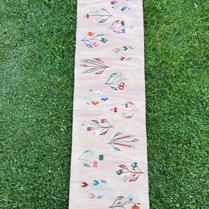 May include: A long pink rug with a floral pattern. The rug has a fringe on both ends and features colorful flowers in shades of red, blue, green, and yellow.