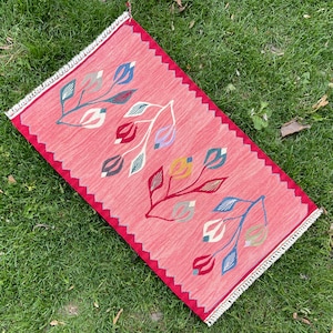 May include: A red rug with a white fringe border features a repeating pattern of stylized floral designs in shades of blue, green, and yellow.