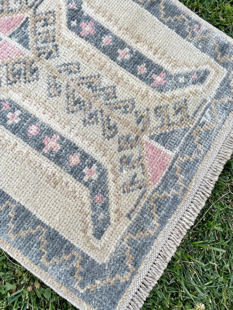 Puede incluir: Una alfombra de color crema con un patr&oacute;n geom&eacute;trico azul y rosa. La alfombra tiene un borde de flecos.