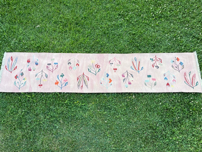 May include: A pink runner rug with a floral pattern. The rug has a fringe border and features a repeating design of stylized flowers in shades of pink, blue, green, and red.