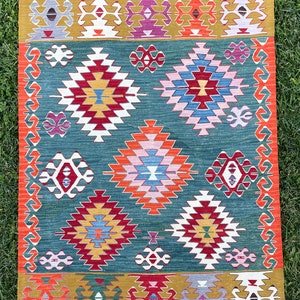 May include: A traditional Turkish kilim rug with a geometric pattern in shades of red, blue, green, yellow, and white. The rug has a fringed border.