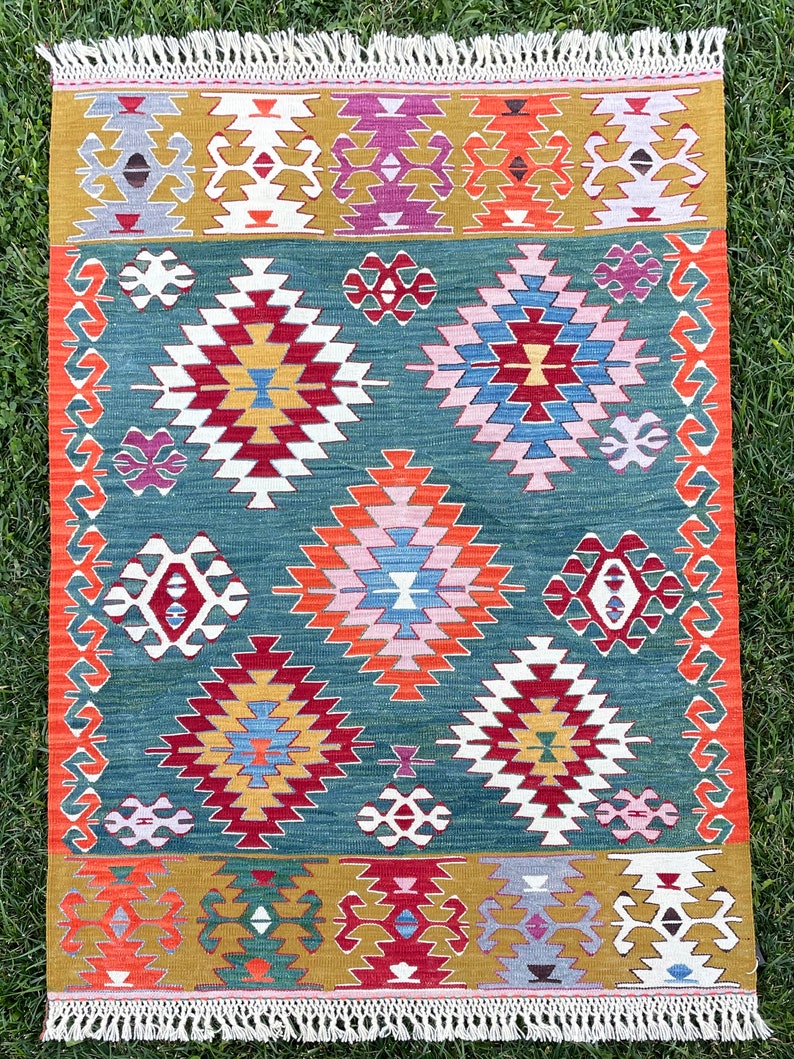 May include: A traditional Turkish kilim rug with a geometric pattern in shades of red, blue, green, yellow, and white. The rug has a fringed border.