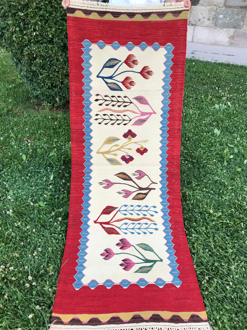 May include: A long, narrow rug with a red background and a white center panel. The center panel features a repeating pattern of stylized flowers in shades of pink, blue, green, and yellow.