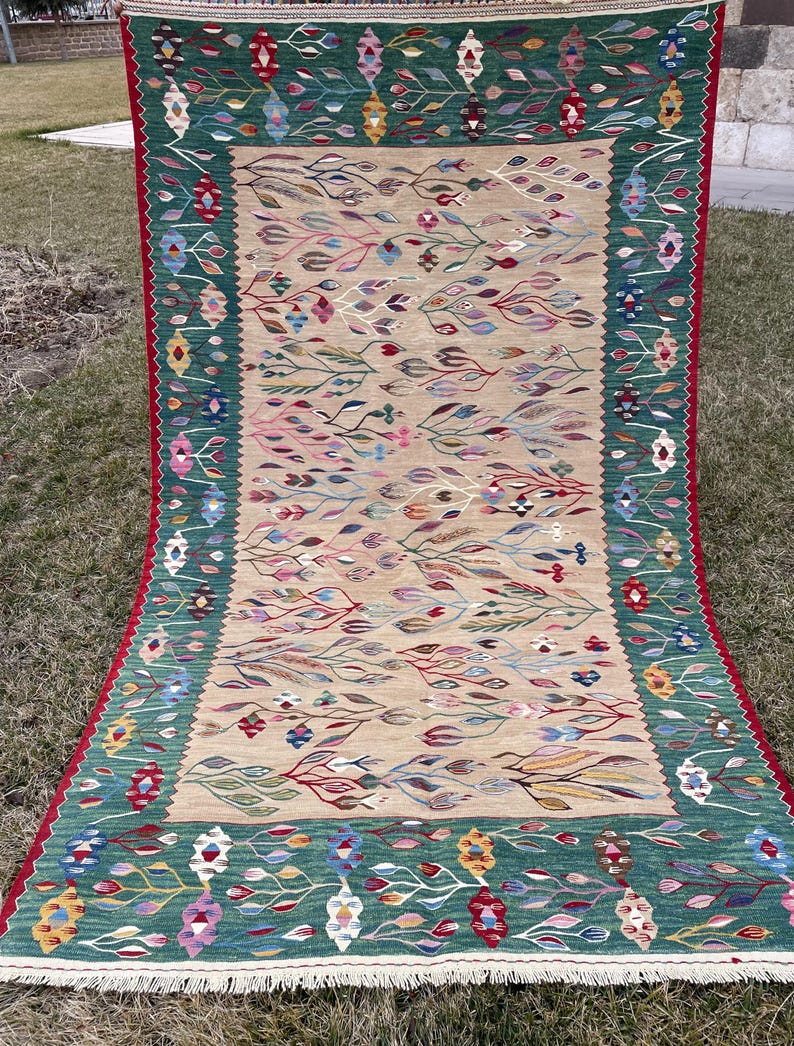 May include: A traditional Turkish rug with a green and beige colour scheme. The rug features a floral pattern with intricate details. The rug has a fringed border.