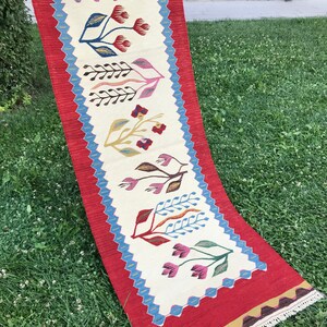 May include: A long, narrow rug with a red border and a white center. The center features a repeating pattern of stylized flowers in shades of pink, blue, yellow, and red.