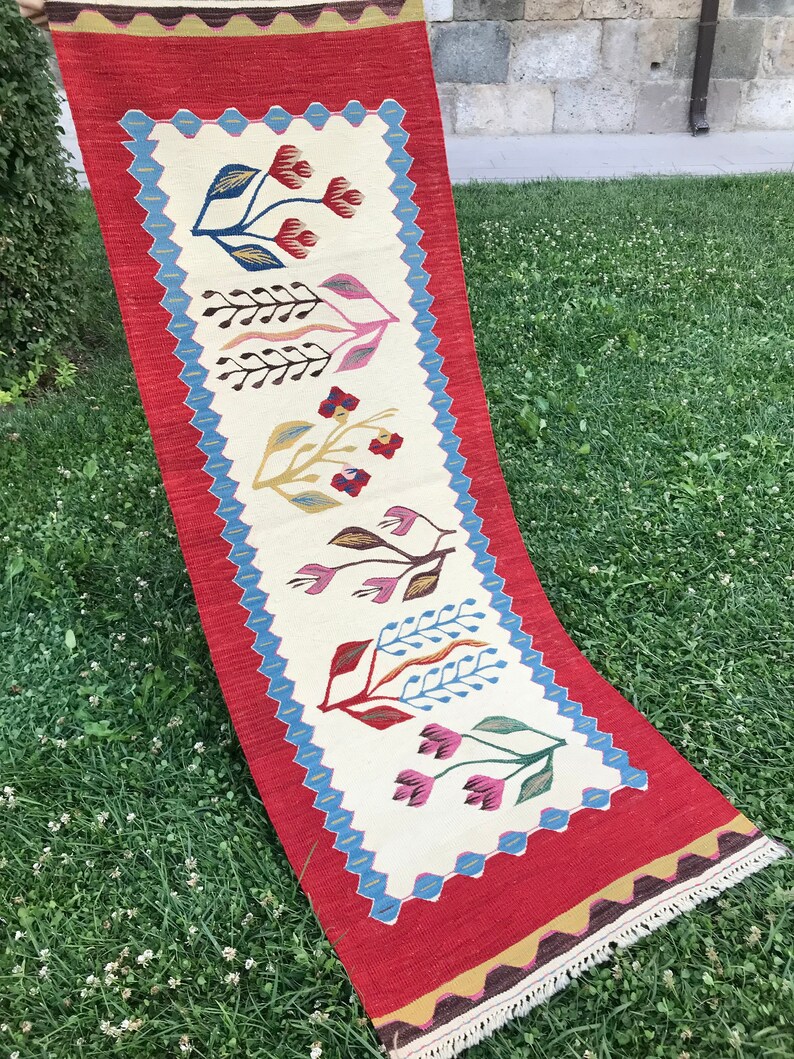 May include: A long, narrow rug with a red border and a white center. The center features a repeating pattern of stylized flowers in shades of pink, blue, yellow, and red.