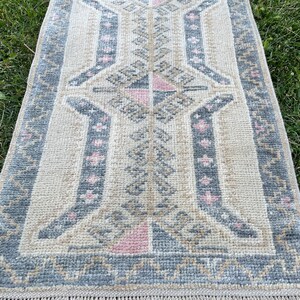 Puede incluir: Una alfombra beige y gris con patrones y acentos rosados. La alfombra tiene un borde de flecos y un dise&ntilde;o geom&eacute;trico.