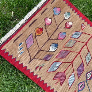 May include: A brown rug with a geometric pattern of colorful leaves and flowers. The rug has a red border with a fringe.