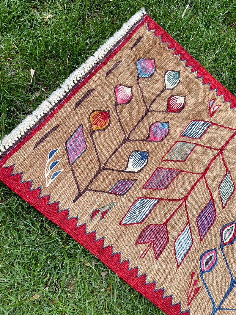 May include: A brown rug with a geometric pattern of colorful leaves and flowers. The rug has a red border with a fringe.