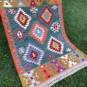 May include: A colourful rug with a geometric pattern in shades of red, orange, yellow, green, blue, and white. The rug has a fringe border and is made of wool.
