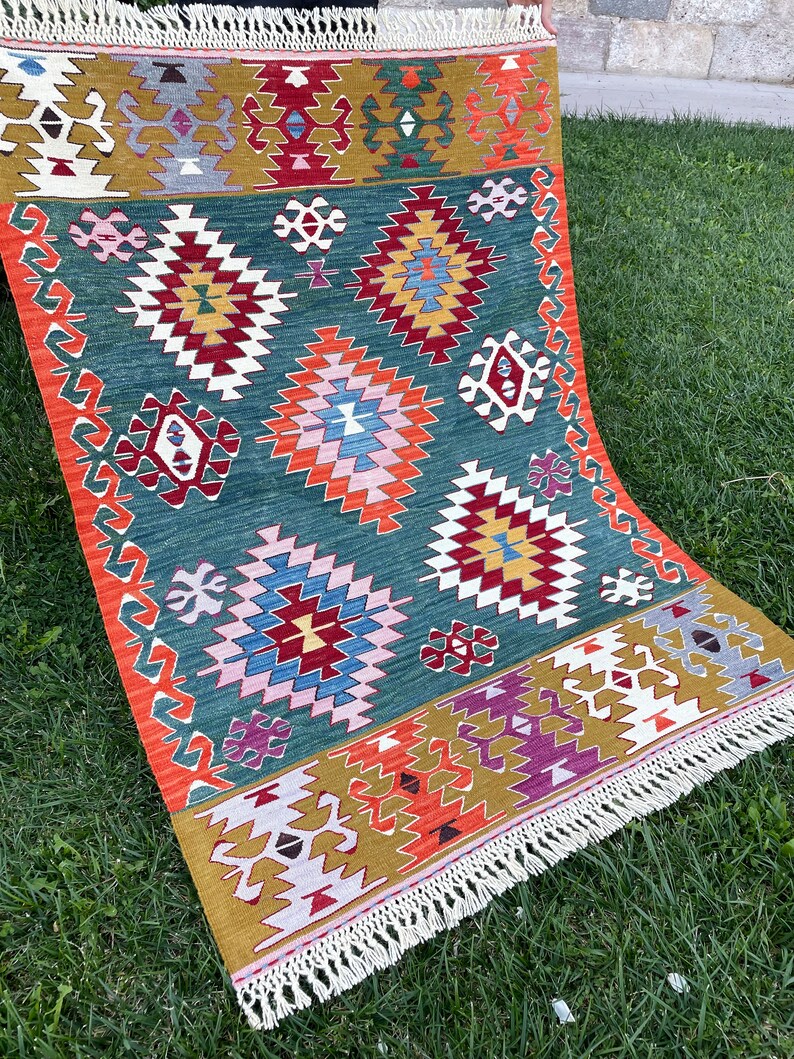 May include: A colourful rug with a geometric pattern in shades of red, orange, yellow, green, blue, and white. The rug has a fringe border and is made of wool.