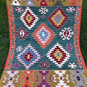 May include: A traditional Turkish rug with a green background and intricate geometric patterns in shades of red, orange, yellow, blue, white, and purple. The rug has a fringe border.