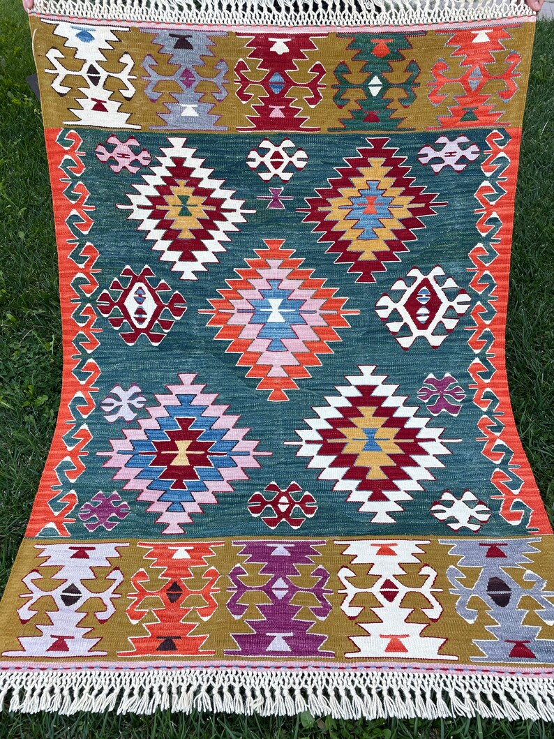 May include: A traditional Turkish rug with a green background and intricate geometric patterns in shades of red, orange, yellow, blue, white, and purple. The rug has a fringe border.