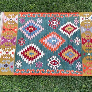 May include: A colourful rug with a geometric pattern in shades of red, orange, yellow, green, blue, and purple. The rug has a fringed border.