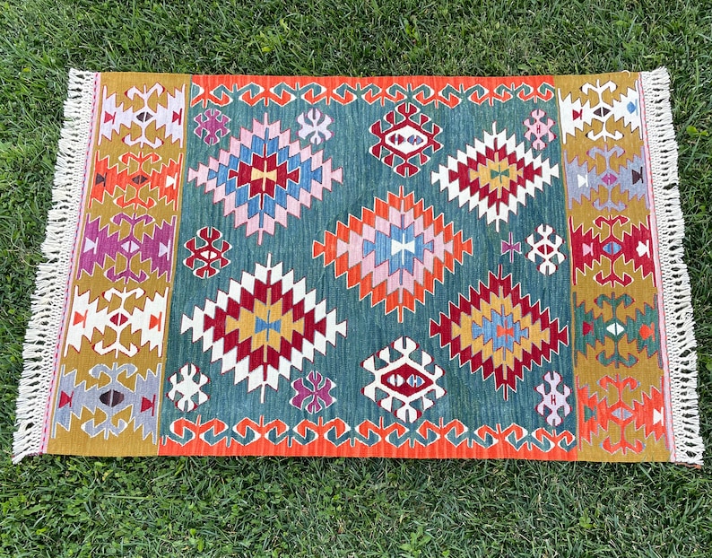 May include: A colourful rug with a geometric pattern in shades of red, orange, yellow, green, blue, and purple. The rug has a fringed border.
