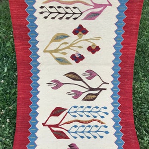 May include: A white runner rug with a red and blue border. The rug features a repeating pattern of stylized flowers in shades of pink, green, brown, and blue.