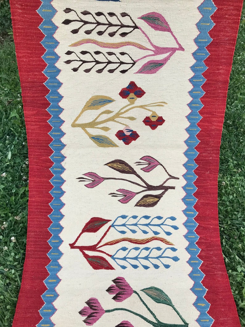 May include: A white runner rug with a red and blue border. The rug features a repeating pattern of stylized flowers in shades of pink, green, brown, and blue.